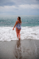 A plump woman in a bathing suit enters the water during the surf. Alone on the beach, Gray sky in the clouds, swimming in winter.