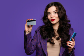Elegant woman in business suit holds plastic credit card and smartphone on purple background indoor. Cashless, contactless payment, money transfer and people concept. Modern digital technology
