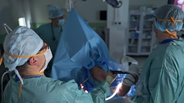 Chief surgeon holds and moves device input into patient's body. Medical team standing around the patient. Blurred backdrop.
