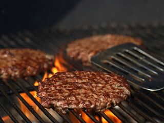 Freshly grilled beef burgers on grill grate with flames and stainless steel spatula in background