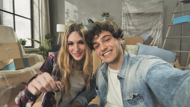 young couple taking selfie in new apartment showing house key phone camera point of view,happy couple sits on the floor of new flat with cardboard boxes belongings,moving to new home concept 4k