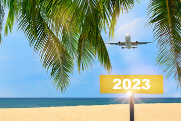 New year 2023 written on yellow sign and commercial plane flying on blue sky with coconut palm...