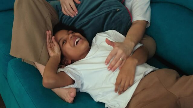 Caucasian mother tickles her African-American daughter at home on the couch,having fun.Diverse people.Time together.Slow motion.