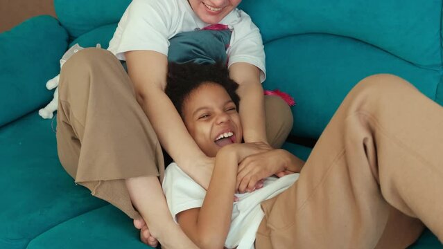 Caucasian mother tickles her African-American daughter at home on the couch,having fun.Diverse people.Time together.Slow motion.
