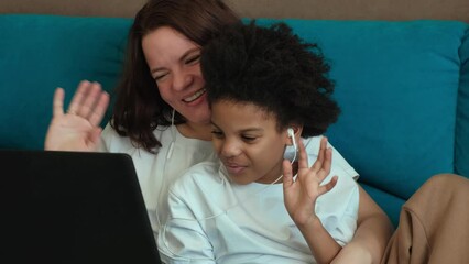 Caucasian mom and an African-American daughter are sitting in an embrace at home on the couch and using a laptop with headphones.Diverse people.Time together.Mother's day.Generation Z.Slow motion. - Powered by Adobe
