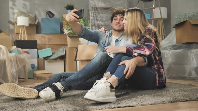 young couple moves to new apartment taking selfie showing house key,happy couple sitting on the floor in living room with belongings cardboards boxes show home key video call with family or friends 4k