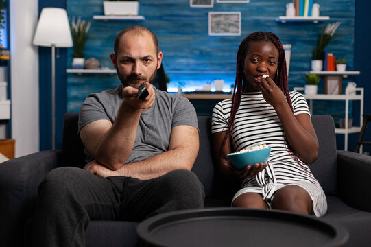 Caucasian man and african american woman watching TV while sharing a bowl of popcorn sitting on sofa at home. Stressed husband angrily switching television programs looking for a movie.