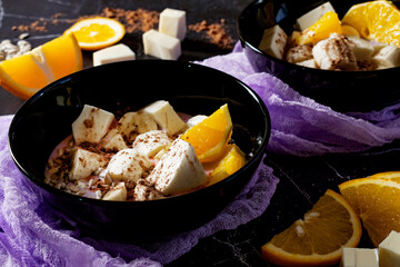 Fruit dessert in blueberry yogurt with bananas, oranges, white chocolate, seeds and sprinkled with cocoa powder, in black plates, ingredients and a lilac textured thin napkin are scattered nearby