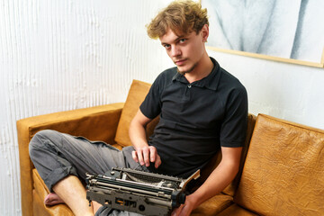 A young European guy is sitting on a sofa with a typewriter in his hands. Image in retro style. Dreamy and romantic young man relaxing in his spare time.