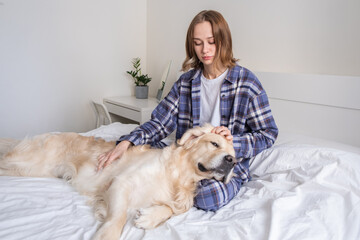 A young woman is happily spending time with her purebred dog. Girl scratching her golden retriever in her bedroom on the bed. Concept: love of animals, friendship, authenticity, happiness, pets