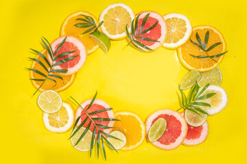 Colorful flatlay of citrus fruit slices and tropical palm leaves. Orange, grapefruit, lemon, lime bright high-colored slices on yellow background. Summer holiday background top view