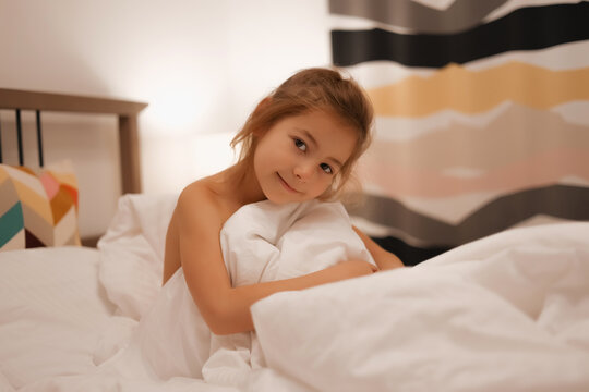 A European girl has woken up and is lying in bed with white pastel linen. Early in the morning, the child wakes up in the garden or school. Bright interior, striped pillows