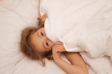 A European girl has woken up and is lying in bed with white pastel linen. The child has woken up and looks out from under the blanket, only his eyes are visible