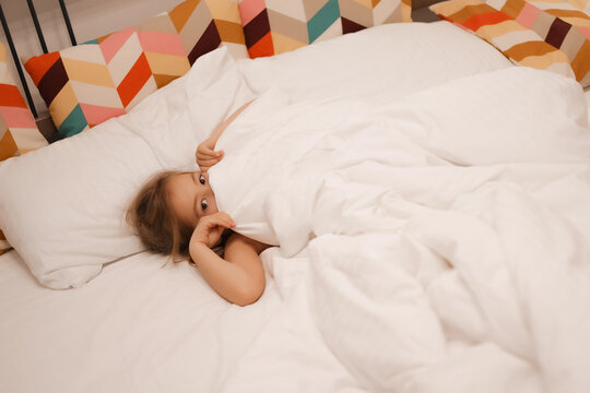 A European girl has woken up and is lying in bed with white pastel linen. The child has woken up and looks out from under the blanket, only his eyes are visible