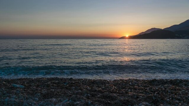 Time lapse of the sun setting below the horizon. View from the pebble beach to the sea. Summer landscape sunset. Albania. Himare. Ionian Sea.