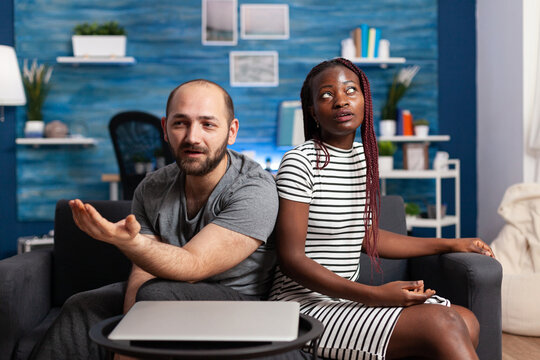 Multiethnic married couple having a dispute on sofa in living room while arguing about financial instability. Irritated partners sitting on couch while having a serious worying conflict
