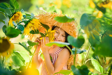 Little pretty girl having fun among field sunflowers. Child hug blooming sunflower. Close up portret. Big fashionable straw hat and striped dress. © Olha
