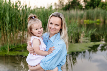 Caucasian Mother with a toddler daughter hugging enjoying outdoors by the river or lake in summer. Happy family portrait. Spending family time together. Single parent