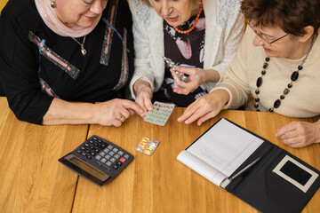 Top view of three absorbed elderly women looking at tablets, sitting at wooden table with notebook with pen, calculator.