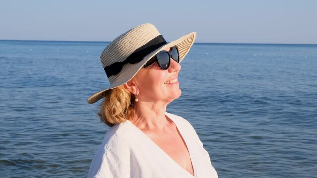 Happy mature woman 50 years old dressed in white dress, straw beach and sunglasses on the beach near the seashore. Vacation concept, relaxation, retirement age