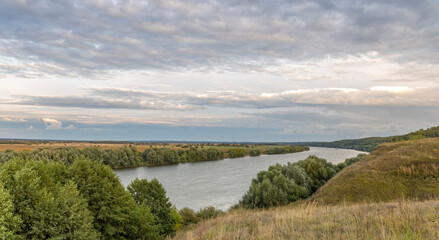 Scenic panoramic view. Green hills, fields and meadows. Sunny summer landscape with a river. Dramatic sky over the horizon. Calm. Clouds over the river.