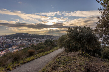 Obraz premium Picturesque landscape of Barcelona from the hill in the early morning. Road in the foreground. Sunbeams through the clouds. Dramatic sky over the city. Autumn in Barcelona, Spain.