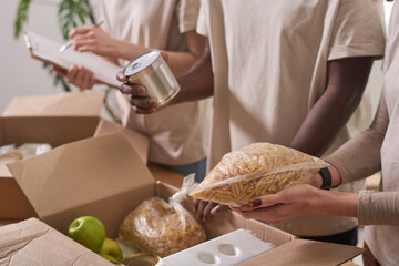 Unrecognizable multi-ethnic volunteers wearing beige T-shirts working together in charity packing food supplies fro donation