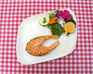A piece of delicious salmon garnished with fresh greens, red radish, onion and lemon on stylish white plate, red chequered tablecloth. Grilled salmon on white plate red and white plaid tablecloth.