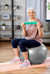 sport, fitness and healthy lifestyle concept - smiling senior woman exercising with resistance band sitting on exercise ball at home