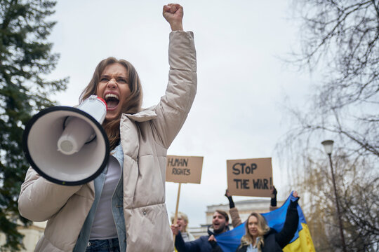 Caucasian woman screaming through megaphone in front and group of young people manifesting against Ukrainian war in the background