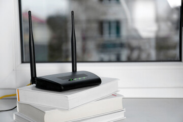 Modern Wi-Fi router on stack of books near window indoors. Space for text