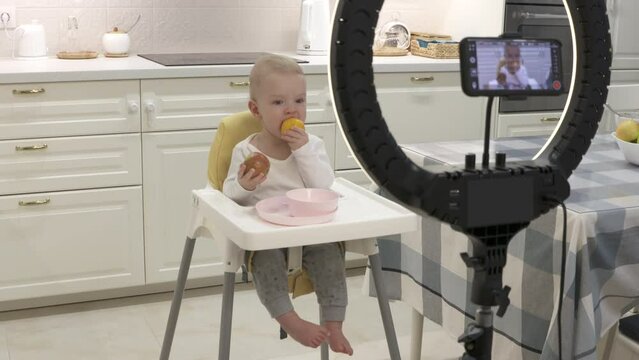 Parents record video on smartphone of kid in high chair at home, recording with mobile phone and ring light photo video of one-year-old baby boy with fruit. High quality 4k footage