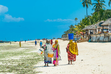 Zanzibar, Tanzania - December 19, 2021: Local women walk by the white sand beach, Zanzibar, Tanzania - obrazy, fototapety, plakaty