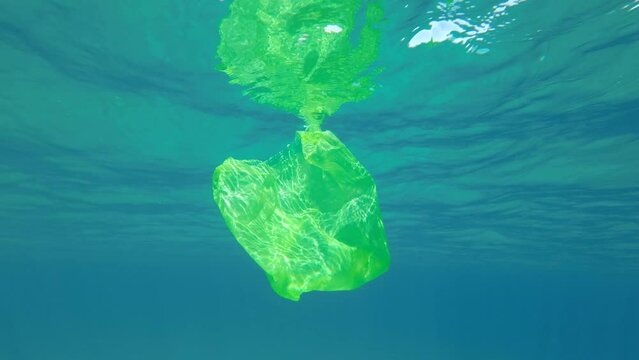 Slow motion, Yellow plastic bag swims underwater reflecting on the surface of the blue water in sun lights. Discarded plastic bag slowly drifting under surface of water in sun light. Plastic pollution