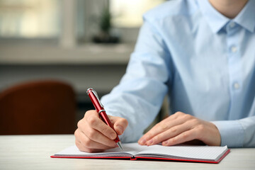 Woman writing with pen in notebook at table indoors, closeup. Space for text