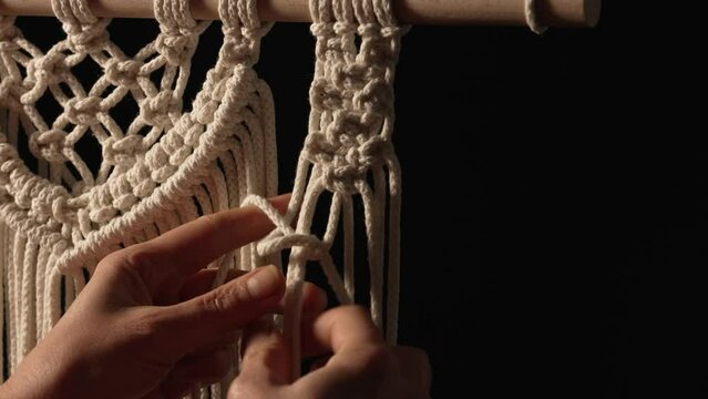 Woman weaves pattern of macrame threads with her own hands for decoration or home decor. Woman's hands tying knots on ropes in a creative workshop on black background. Handmade. Close up. Slow motion.