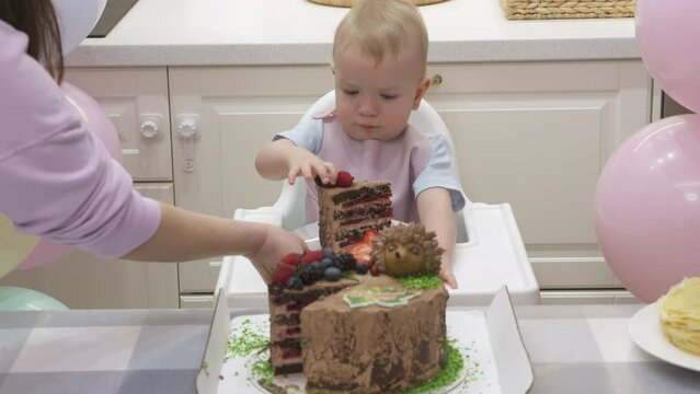 Baby celebrate first birthday holiday, mother cuts pieces of birthday cake, one year old kid against colorful balloons at his first birthday party. High quality 4k footage