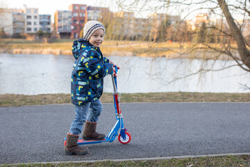 Cute toddler boy, riding a scooter in a park on a cold sunny day