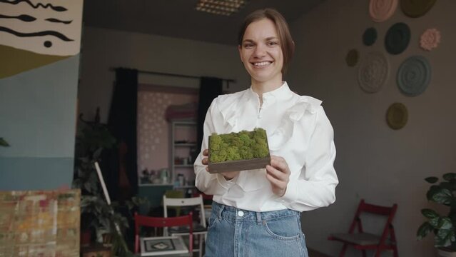 A tender young girl demonstrates a picture of stabilized moss in the original interior of a room with many indoor plants. Slow motion