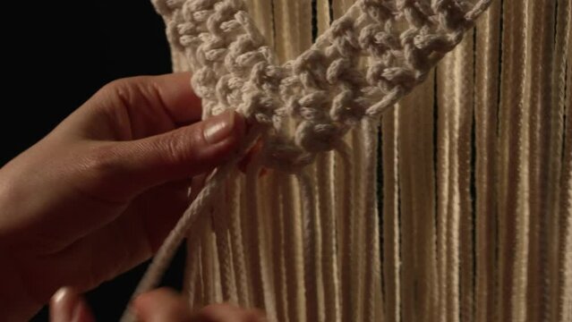 Women's hands weave a macrame pattern in creative workshop on black background. Woman ties knots on cotton threads to create lace. The base knots of macrame. Craft of rope. Close up. Slow motion.