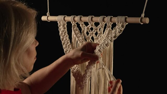 Weaving macrame pattern from natural white cotton threads. Women's hands knit knots creating lace ornament on black background. Creative process in workshop, textile crafts. Close up. Slow motion.