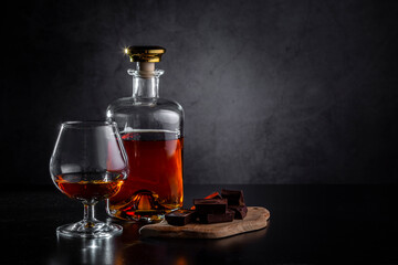 A glass of cognac, cognac in a bottle and pieces of chocolate on a dark background. Copy space
