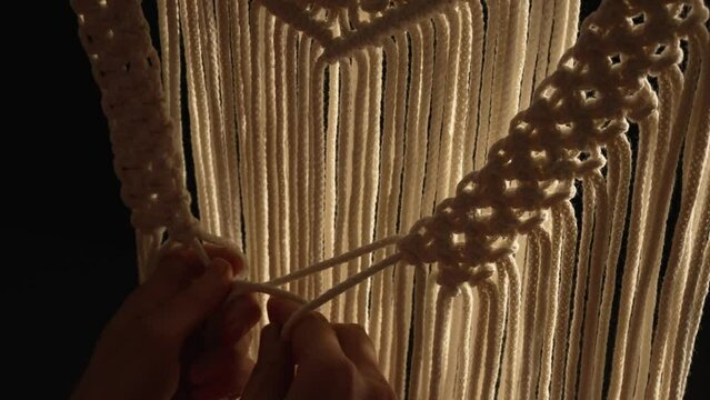 Weaving macrame pattern from natural white cotton threads. Women's hands knit knots creating lace ornament on black background. Creative process in workshop, textile crafts. Close up. Slow motion.