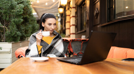 A girl in a cafe on the street wrapped in a plaid, enjoying a sunny warm autumn day. Cup of coffee tea in hands Young brunette working behind a laptop.