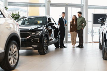 Confident salesman showing new car to young Caucasian couple at auto dealership