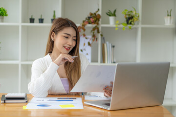 Beautiful Asian woman holding a graph document working on a laptop computer at the office. businesswoman looking at financial statistics data analysis chart graph profit growth business idea