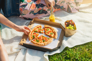 Two women eating fresh pizza from a paper box sitting on a grass on a sunny day having picnic drinking orange juice and having grapes