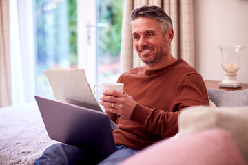 Mature Man At Home Sitting On Sofa With Laptop Looking Through Personal Finance Documents