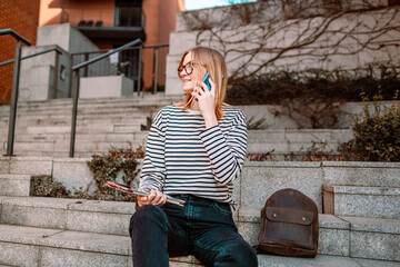 Stylish woman with blonde hair and eyeglasses talking on the phone on the street in warm good day. Communication technology.