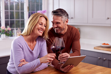Mature Couple At Home With Digital Tablet Celebrating Booking Holiday Or Purchase With Red Wine